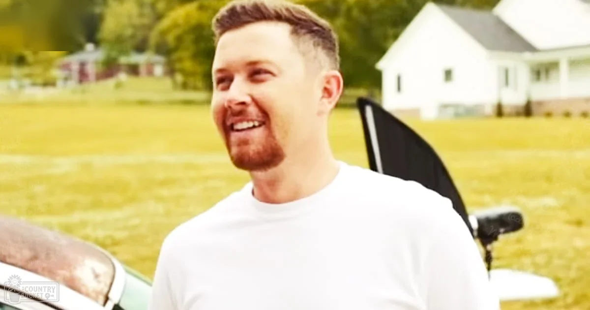 A young adult male in a white t-shirt stands outdoors amidst lush greenery and suburban houses, wearing casual attire typical of spring or summer days.