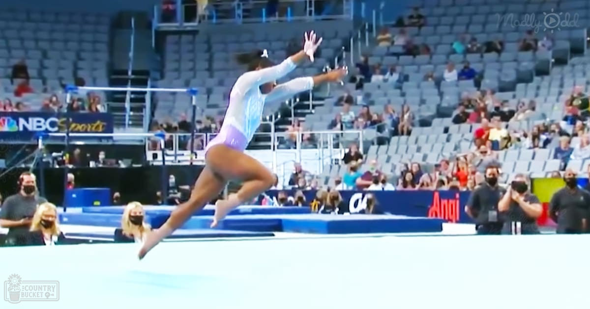 Young female gymnast performing in an indoor sports arena, surrounded by spectators and media; light blue leotard with white and purple accents, barefoot on a bustling gym floor.