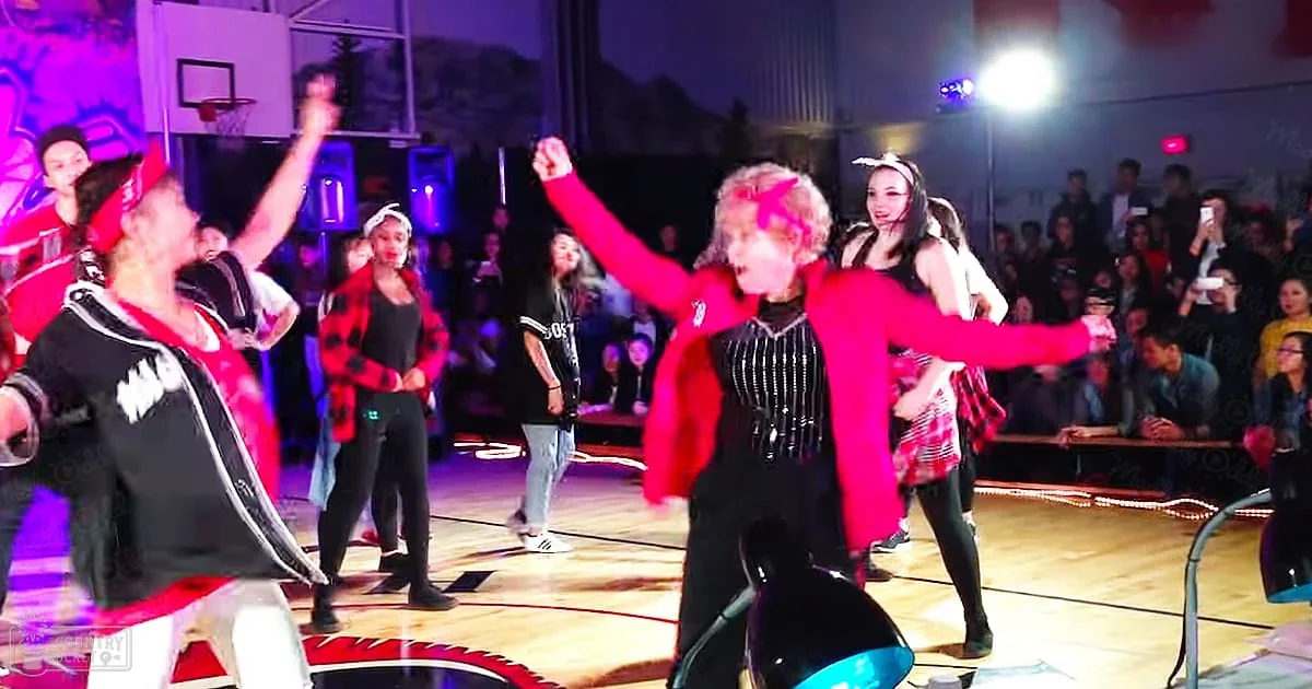 Young adults performing in a school gymnasium with artificial stage lighting, red and black clothing predominant.
