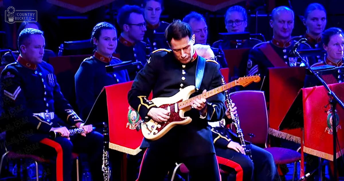 Musicians performing at a formal indoor event with red chairs and stage lighting, focusing on the foreground guitarist in uniform.