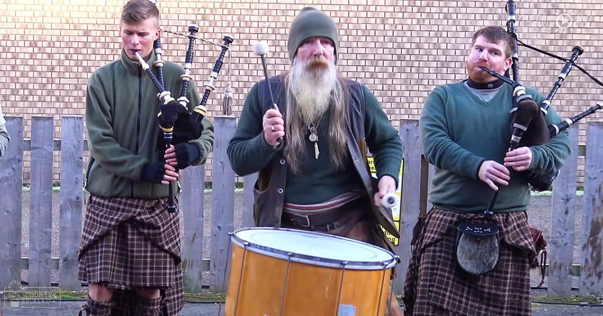 A group of young adult males playing bagpipes and a drum outdoors near a brick wall and wooden fence, with natural light suggesting daytime.