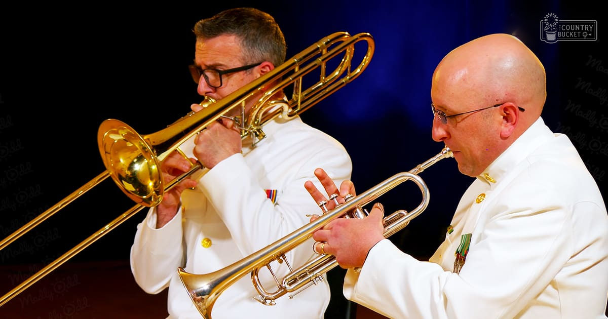Two adult male musicians, likely military or ceremonial attire, playing brass instruments on a stage with dark blue backdrop under professional lighting.