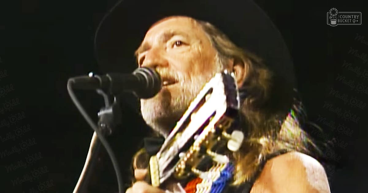 An older adult male singer wearing a dark long-sleeved shirt and ornate necklace performs into a microphone at an indoor concert or music event.
