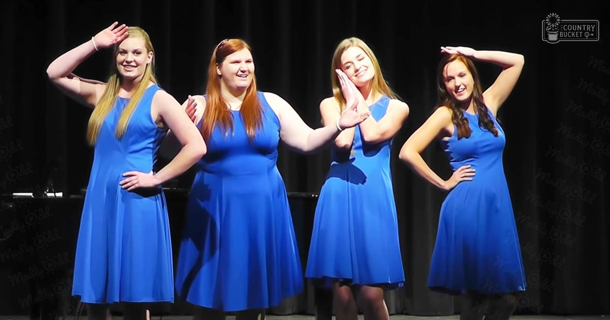 A group of young adult females performing on stage, wearing matching sleeveless blue dresses with empire waists and flared skirts, under artificial lighting in an indoor setting.