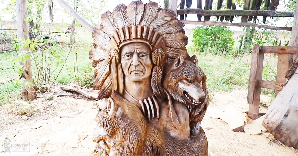A carved wooden statue of an older adult male wearing traditional Native American attire stands outdoors in a rural setting with natural elements like trees and grass.