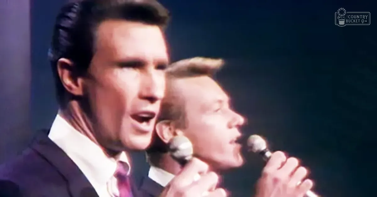 A dimly lit stage with two adult male performers wearing dark suits and white shirts, one holding a microphone against a backdrop of dark curtains.