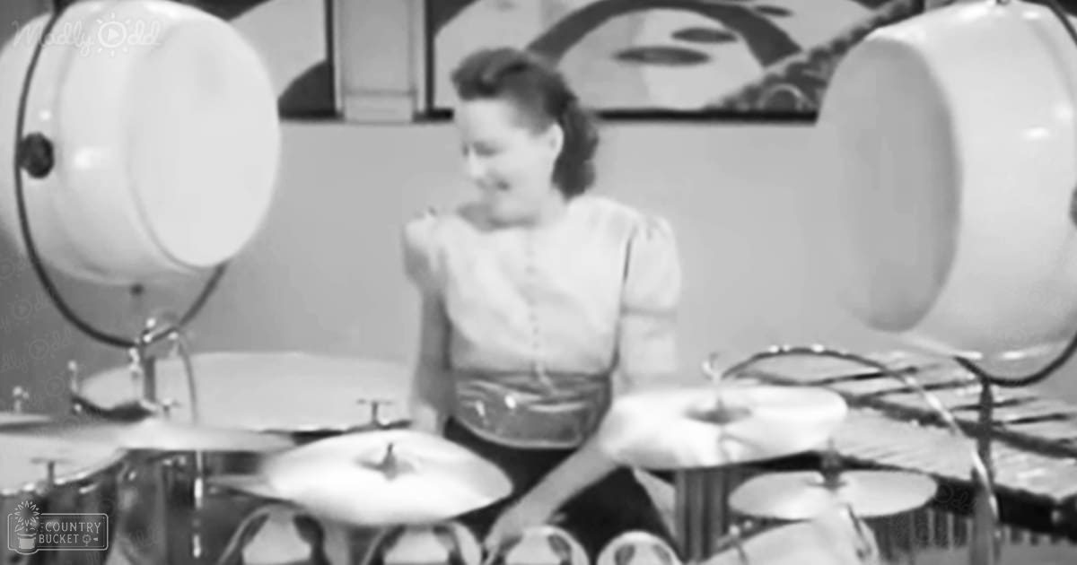 A young adult female playing drums in an indoor studio with a mid-20th-century style, wearing a white vintage-style blouse and surrounded by musical instruments.