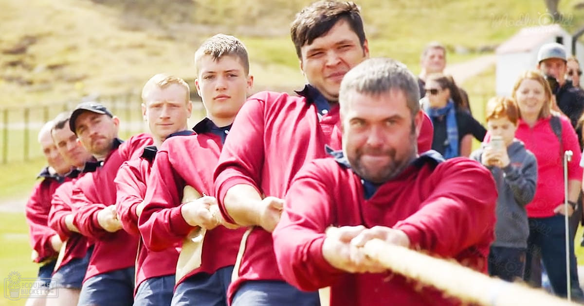 A group of adults participating in a tug-of-war event outdoors on a sunny day, with greenery and grassy hills in the background.