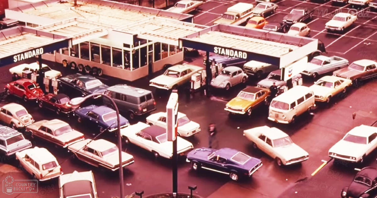 1970s gas station scene with parked cars at pumps, Standard sign visible, muted tones of pink, yellow, and brown, spring or summer season, daytime light, commercial building in background.