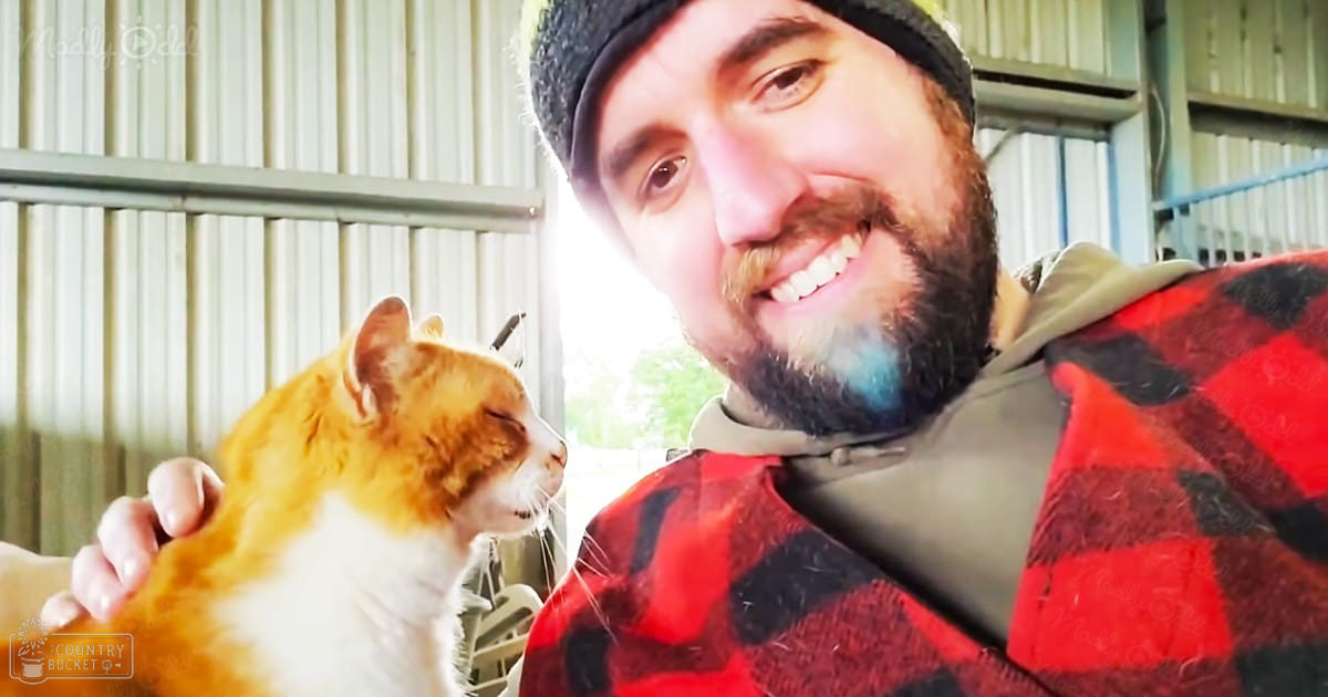 An adult male holding an orange tabby cat in a barn setting during daytime with natural light.