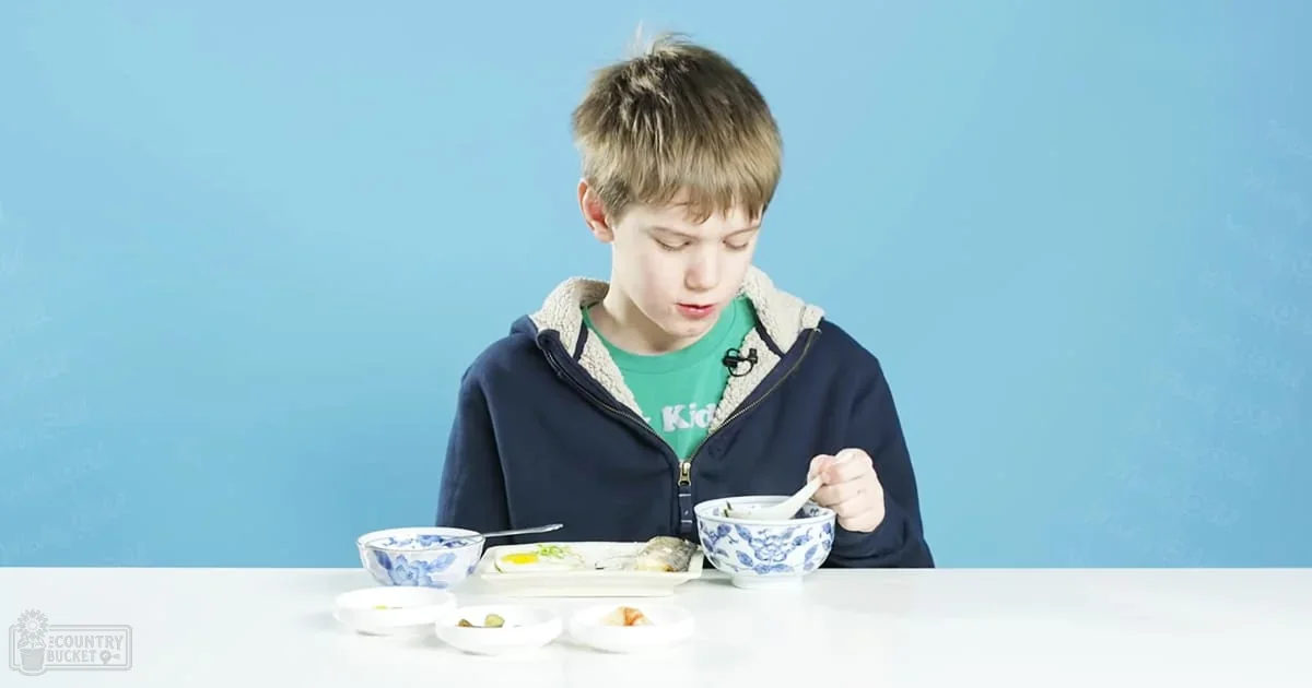 A young adult male child in a casual dark blue zip-up hoodie with a light-colored fleece lining, holding a spoon and wearing a microphone clipped to his hoodie, standing against a plain blue background in an indoor studio setting.