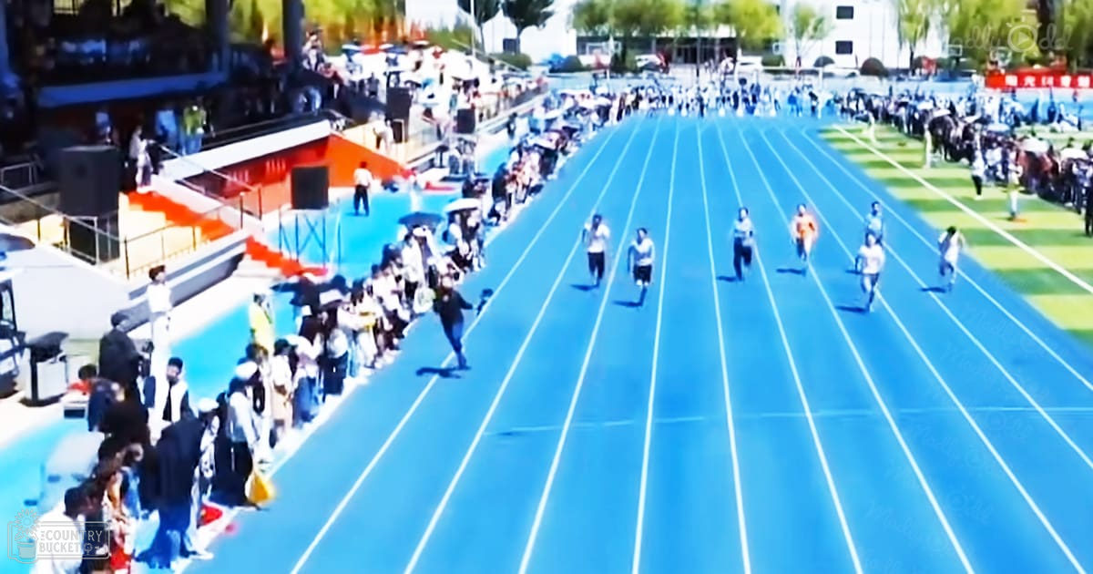 An outdoor stadium with running tracks, bleachers filled with spectators, trees, buildings, and a school or university campus setting during spring or summer with clear daytime weather.