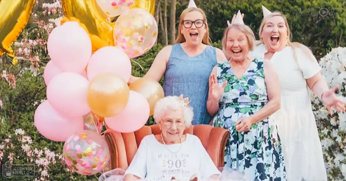 An older adult woman celebrating a milestone birthday with her family outdoors, surrounded by blooming flowers and colorful balloons.