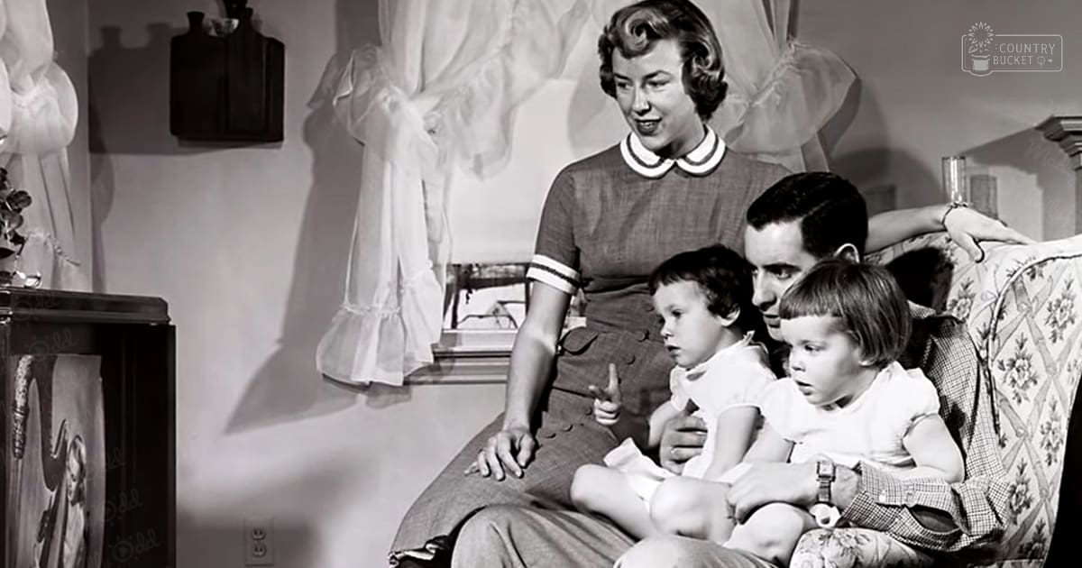 A family scene indoors, with an adult female in a Peter Pan collar dress with ruffled hem, an adult male in a plaid shirt and trousers, two toddlers in white dresses, floral-patterned armchair, dark wooden cabinet, sheer curtains suggesting daytime.