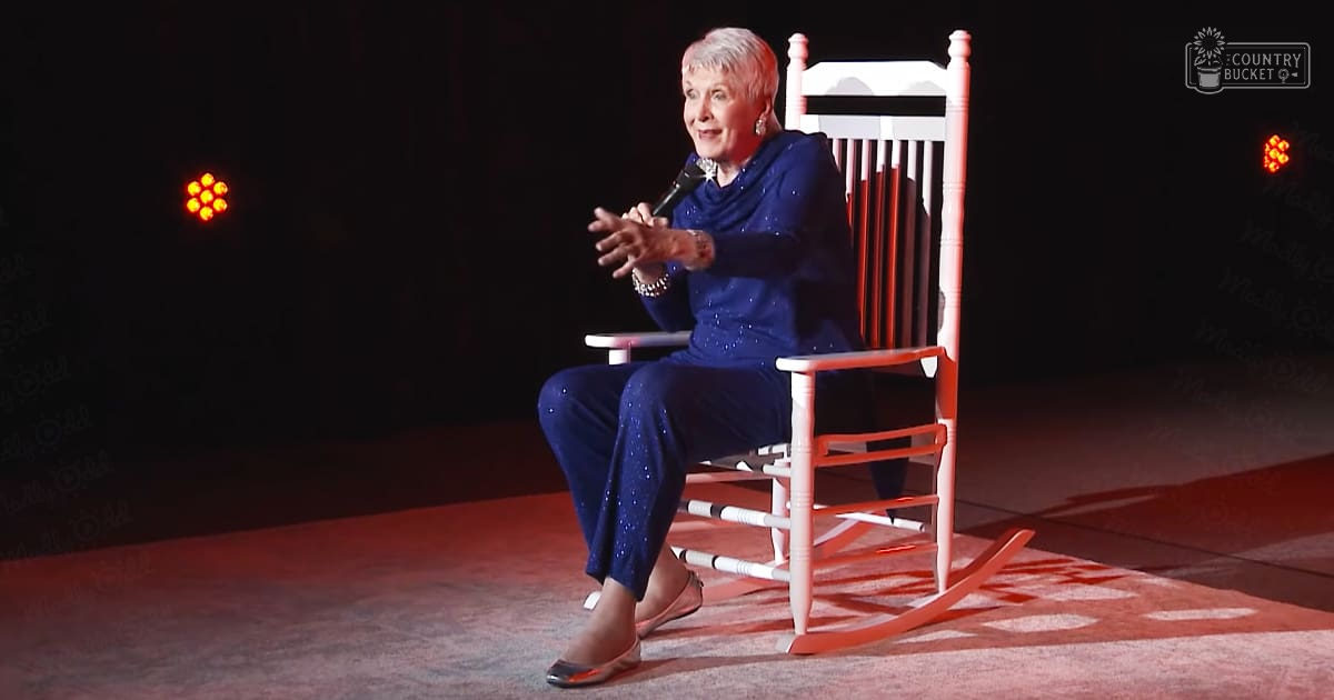 An older adult woman in a blue sequined dress, sitting on a white rocking chair with red accents and holding a microphone, performing indoors under stage lights.