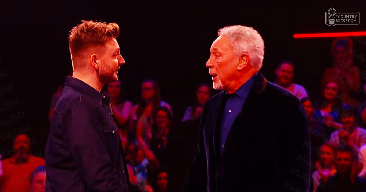 Two adult males, one younger with a dark blue button-up shirt and another older in a dark suit, on an indoor stage or performance area with dim lighting.