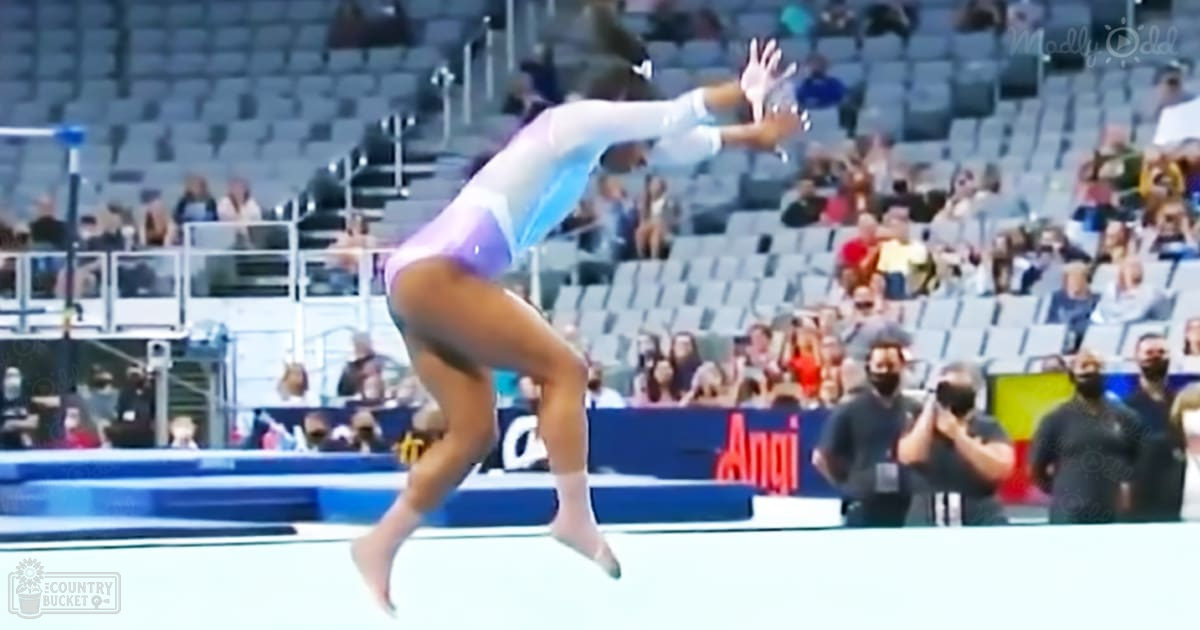 A young adult female gymnast performs at an indoor arena, surrounded by spectators and photographers.