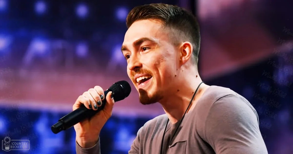A young adult male performs on stage, wearing a relaxed fit long-sleeved gray shirt with crew neck and holding a microphone under purple lighting.