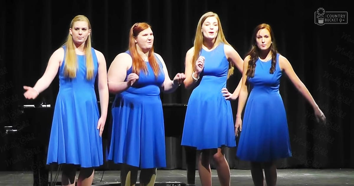 Four young adult women performing on stage in sleeveless blue dresses, with a piano partially visible behind them under artificial lighting.