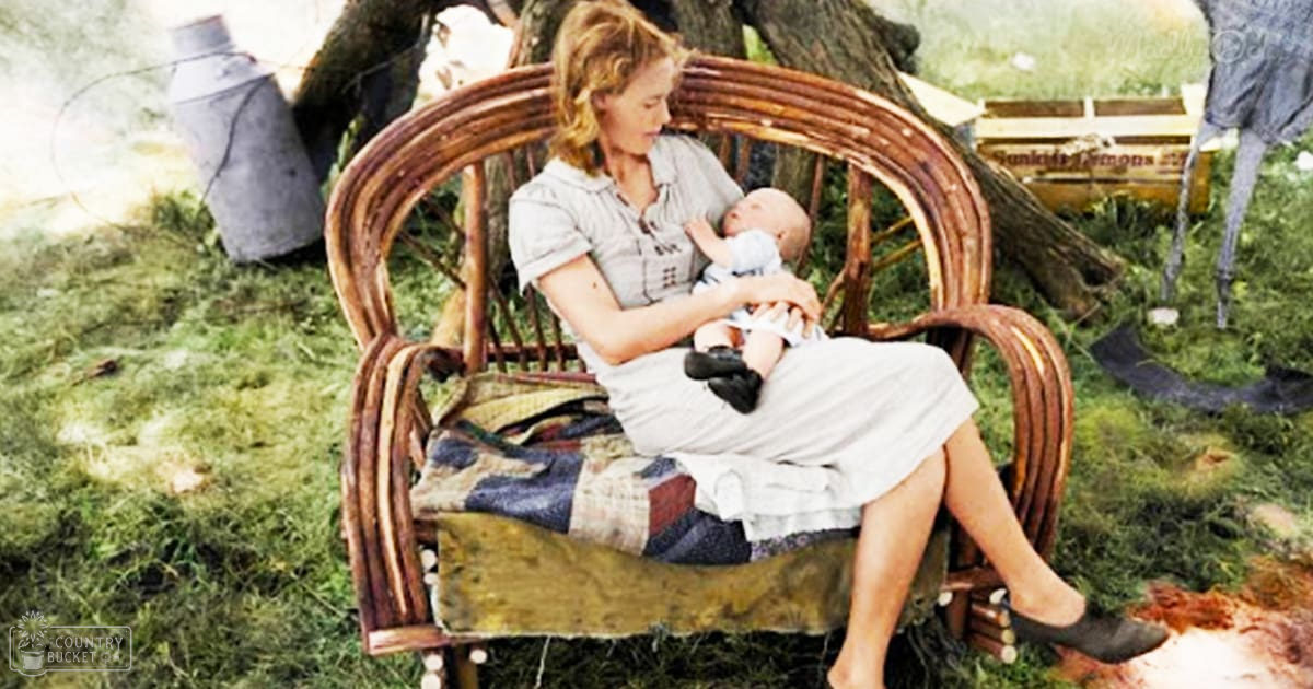 A woman in a light gray dress holds an infant outdoors in a rural setting with rustic furniture and farm items.