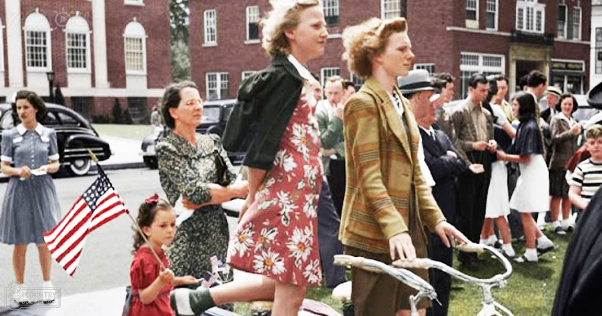A parade scene with a crowd dressed in 1940s or early 1950s attire, with vintage cars and an American flag held by a young girl in front of a brick building.