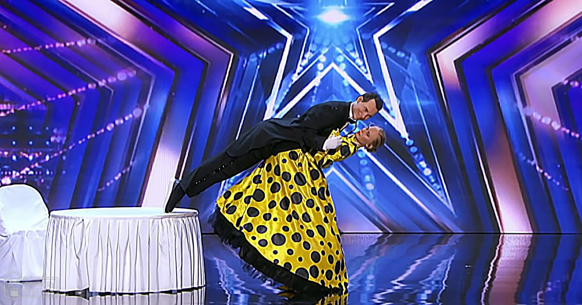 A dance performance scene with a male and female young adult in formal attire, set against an elaborate blue-lit geometric backdrop. A round table with white cloth is partially visible on the left.