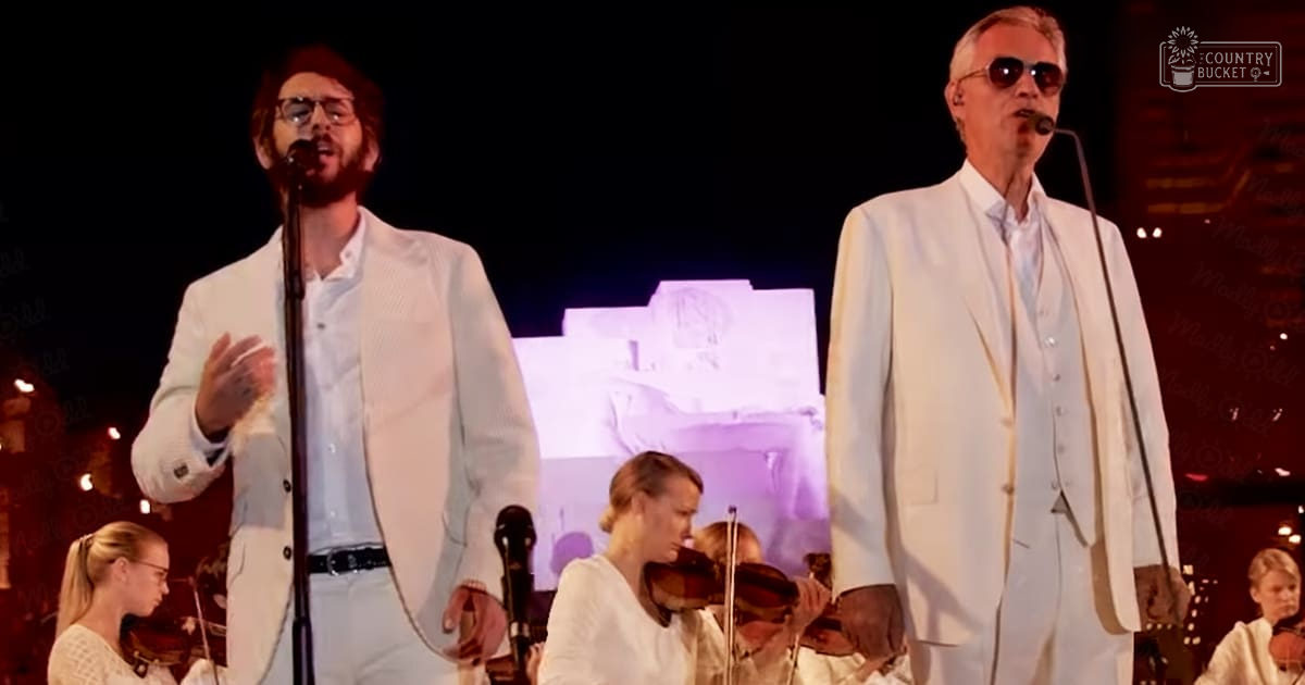 Musical performance with two male singers performing on stage at an indoor concert venue, surrounded by string orchestra musicians.