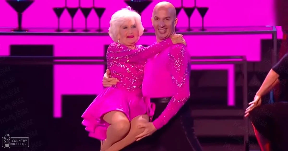 An older adult female dancer wearing a bright pink, sequined dress performs alongside an adult male dancer in a similar outfit on stage at a ballroom event. The setting includes a bar counter with glasses and vibrant pink lighting.