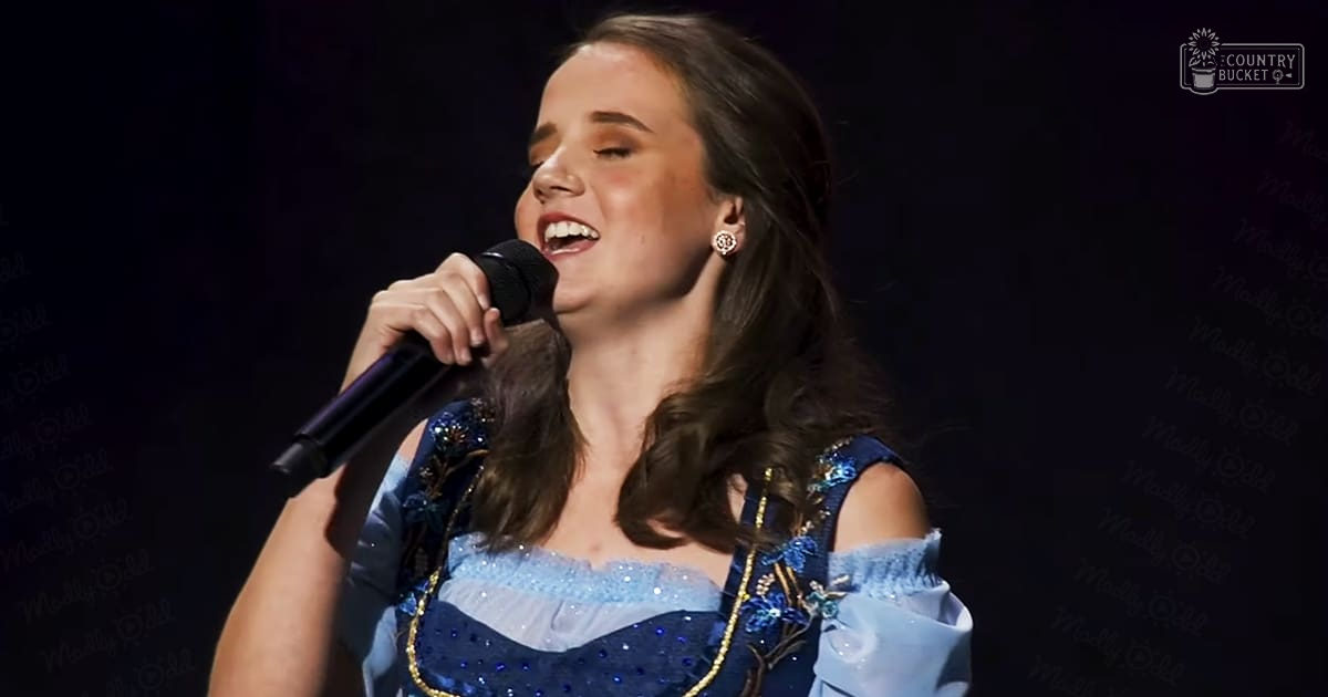 A young adult female in a blue dress with floral embellishments and sequins performs on stage, spotlighted against a dark background.