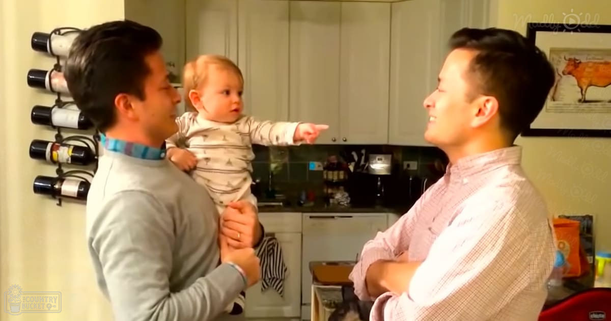 A baby in a white onesie with small triangles, held by an adult male wearing a light gray shirt and a blue and pink striped collar, standing in a kitchen setting with light-colored walls, white cabinetry, and natural daylight.