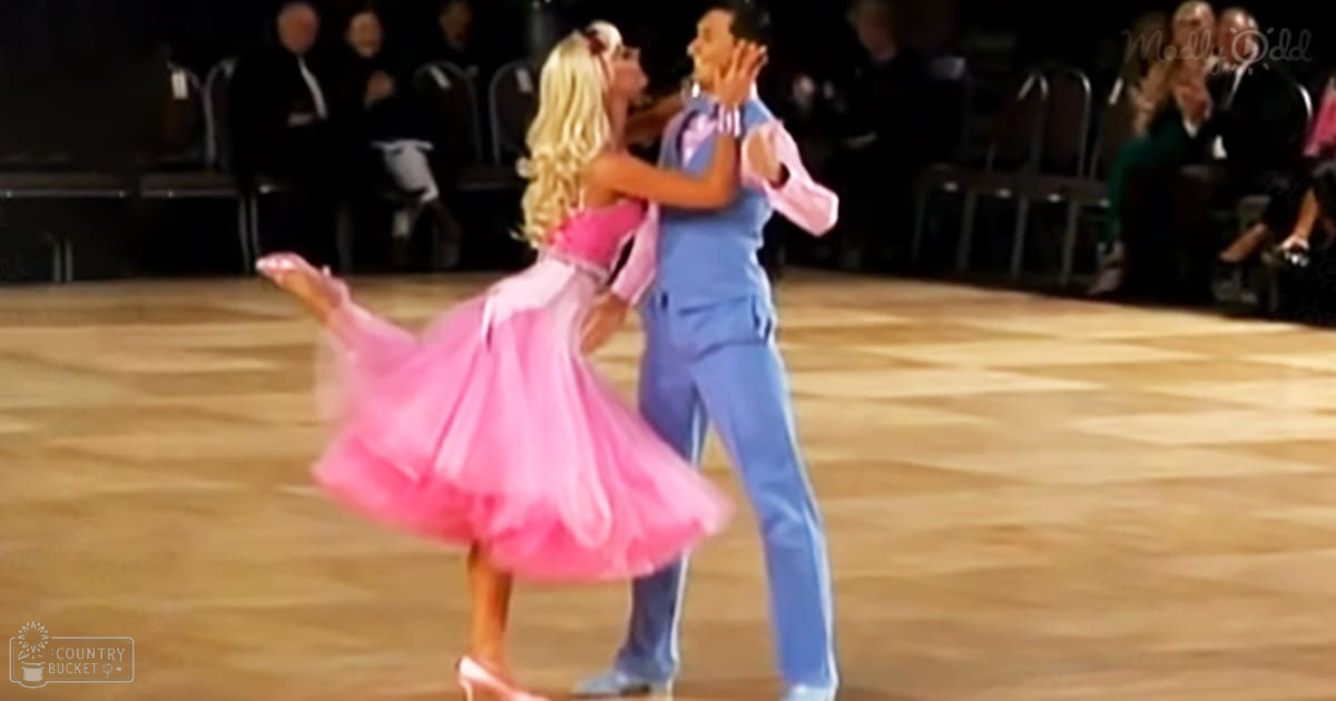 A couple dancing in a formal ballroom setting, the woman wearing a flowing pink dress with a fitted bodice and full skirt, and the man dressed in a blue 1950s-style suit.