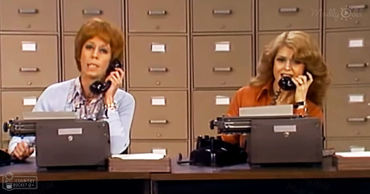 Two women in an office setting with typewriters and filing cabinets, wearing light blue and orange shirts respectively.
