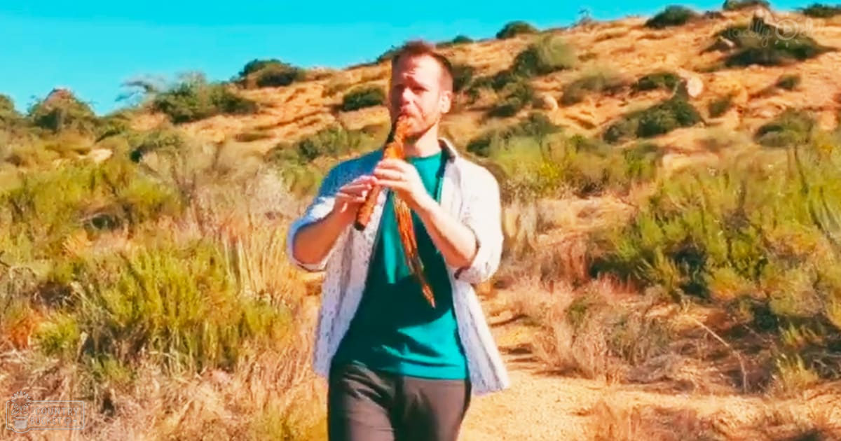 Adult male playing a flute in an open field with dry grass and shrubs under clear blue sky, late summer or early autumn.
