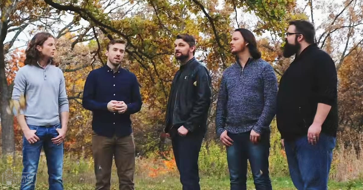 Five young adult men in an autumnal outdoor setting with trees displaying vibrant foliage, wearing casual attire such as button-up shirts and jeans.