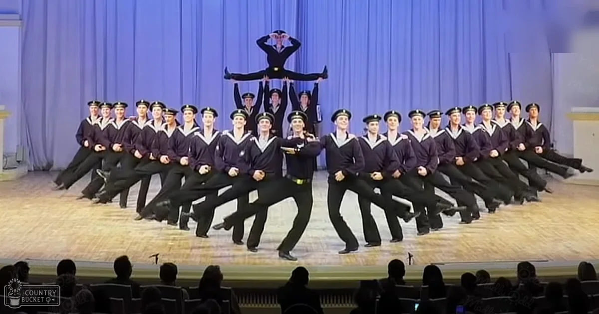 A group of young adult male performers in uniform dark blue sailor-style jackets with white collars and cuffs, black trousers, and dark caps or hats, performing on a stage with wooden floor and blue curtains. An audience is seated in rows of chairs at the bottom edge of the frame.