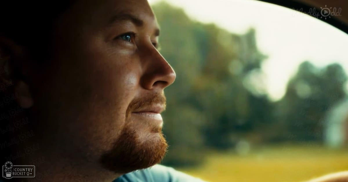 Adult male in a light blue t-shirt, seated inside a vehicle with greenery visible through the window, suggesting an outdoor setting during late afternoon.