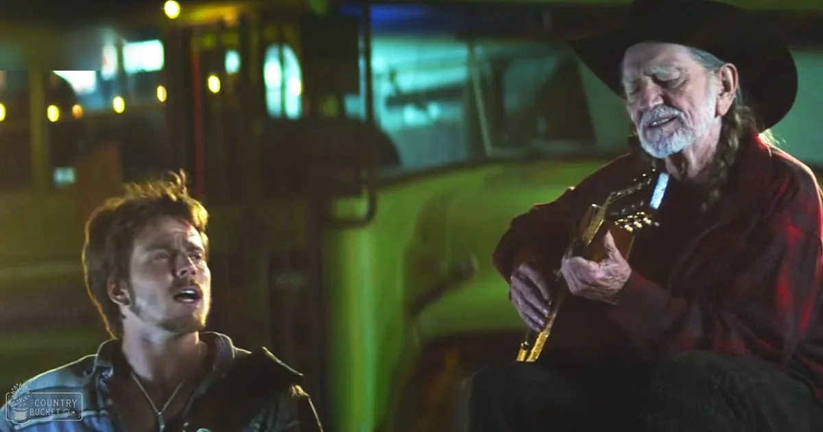Two men playing guitar outdoors at night under artificial light, wearing plaid shirts and a cowboy hat in an urban setting.