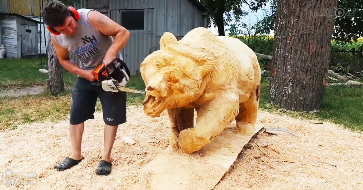 A man in a gray tank top and dark shorts carves a bear with a chainsaw outdoors during summer under natural daylight.