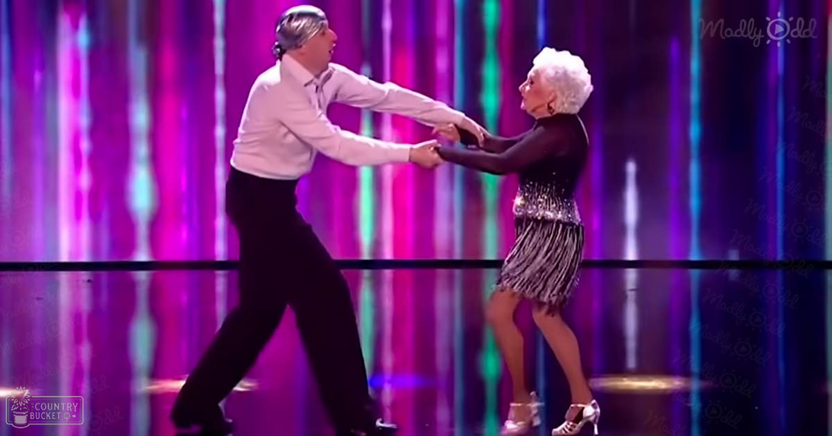 A dance performance scene in a studio or stage setting with vibrant vertical light strips, reflective flooring, and two dancers wearing formal attire. The male dancer wears a white long-sleeved shirt and black pants, while the female dancer is dressed in a black dress with silver fringe details and white high-heeled shoes.