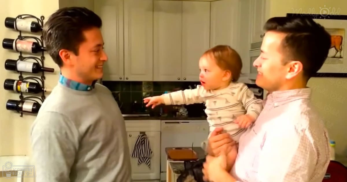 A baby being held by an adult male in a kitchen setting with white cabinets, black countertop, and various household items visible.