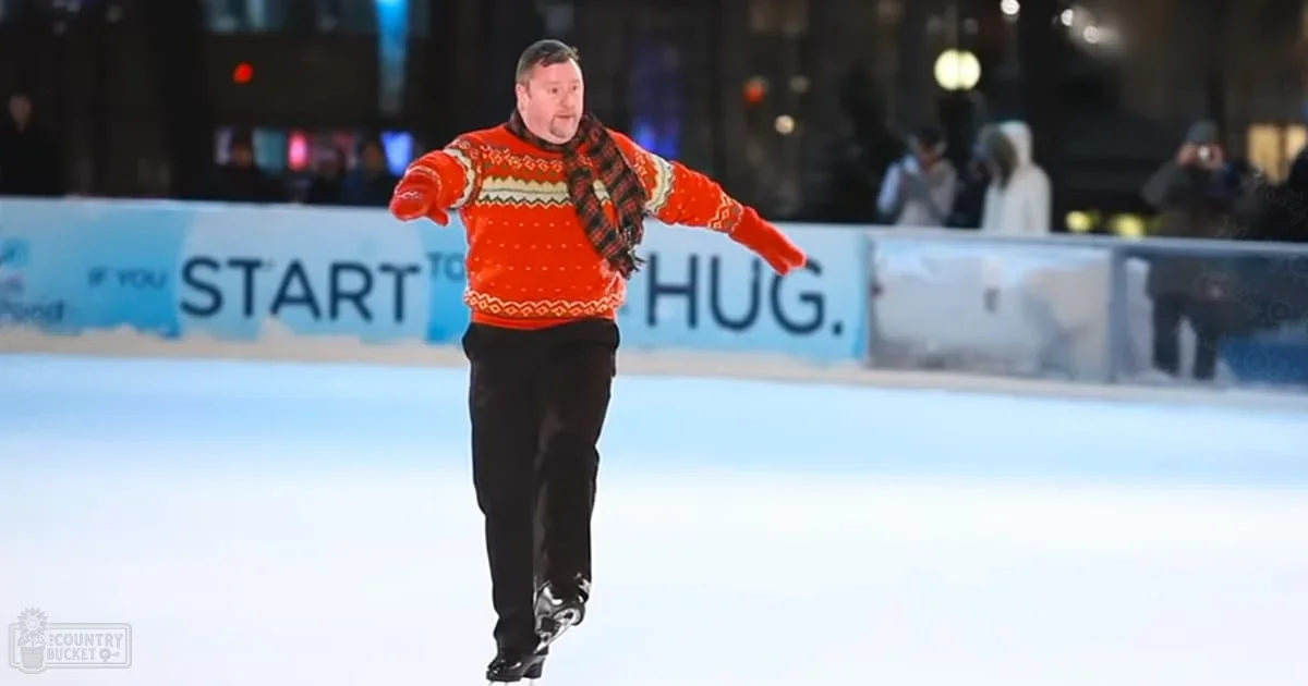 An adult male skating on an outdoor ice rink at night, wearing a red sweater with white and black patterns.