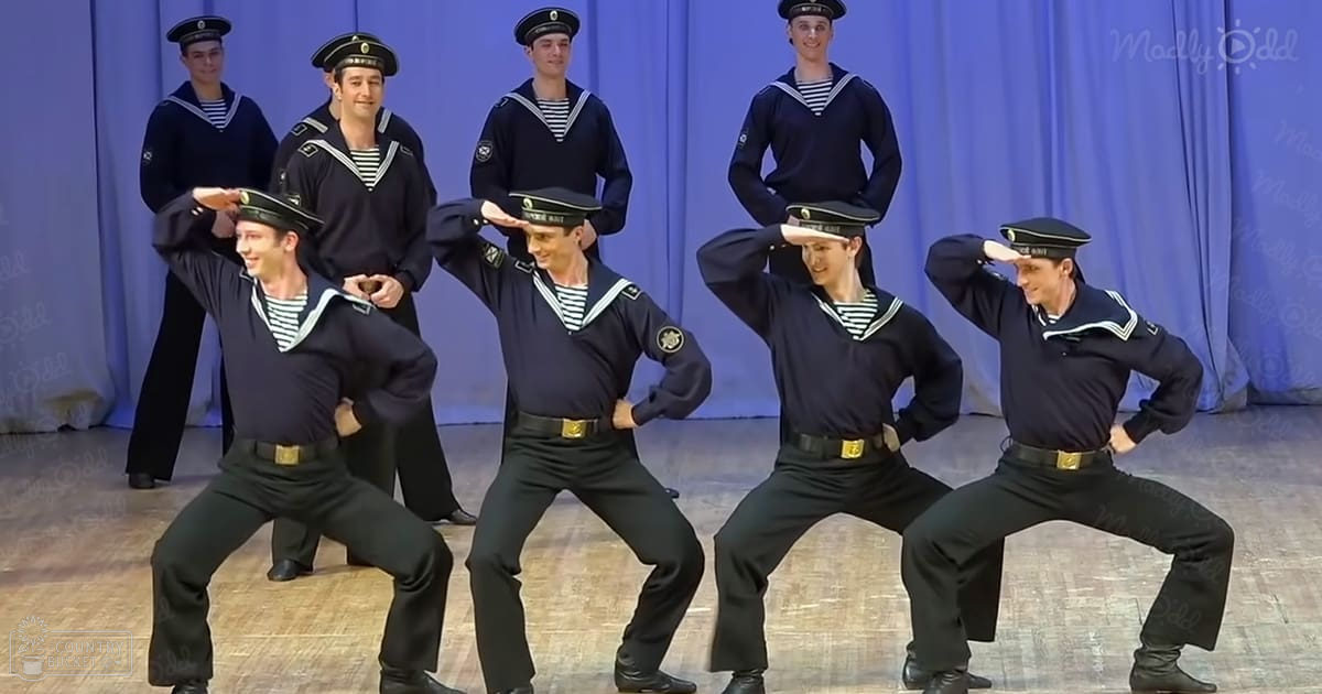 A group of young adult males in a studio setting, wearing sailor-style uniforms with striped shirts underneath, black pants, gold belt buckles, and black shoes or hats with green bands.