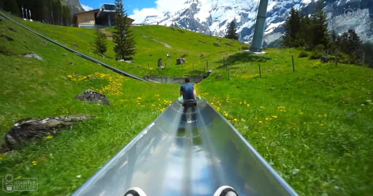 A young adult male slides down an alpine slide in Grindelwald, Switzerland, surrounded by snow-capped mountains and a partly cloudy sky.