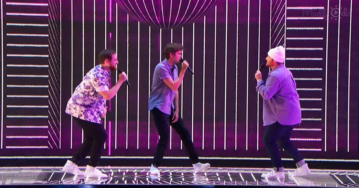 A performance scene with three adult male singers in a modern indoor venue with stage lighting and geometric patterns behind them.