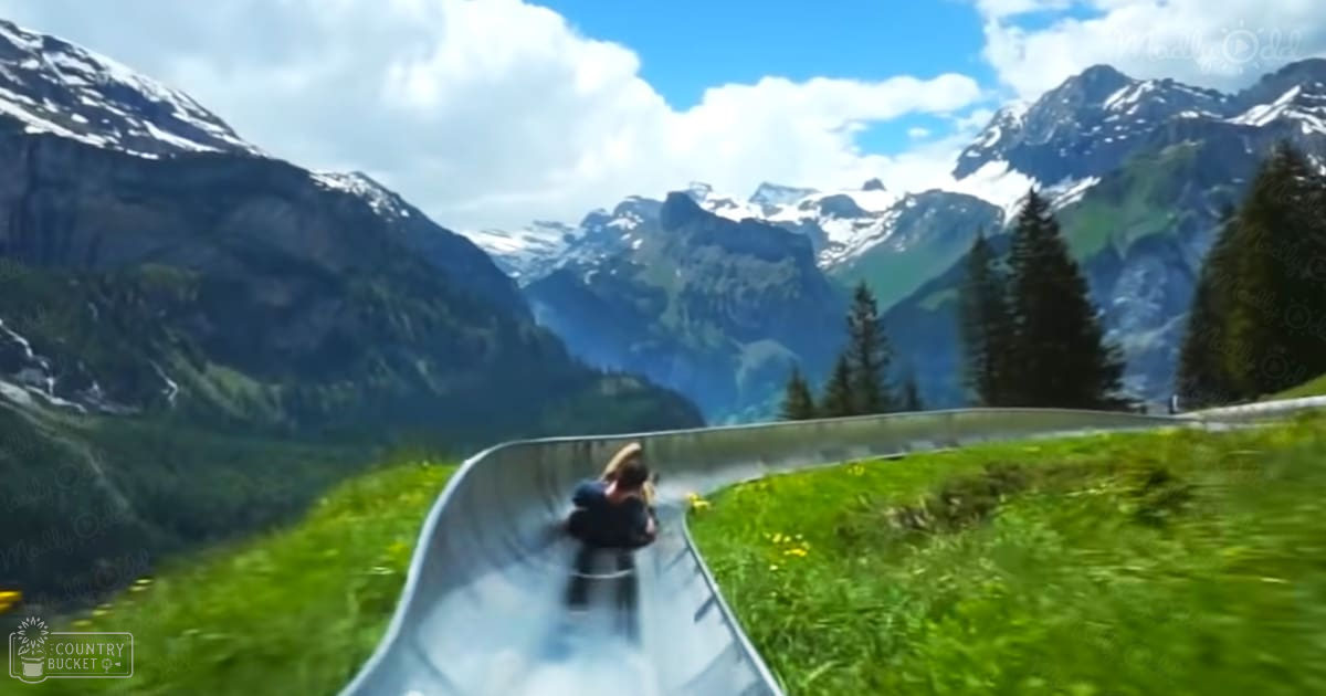 Two children sliding down a hill in a lush green mountainous area with snow-capped peaks in the background during daytime.
