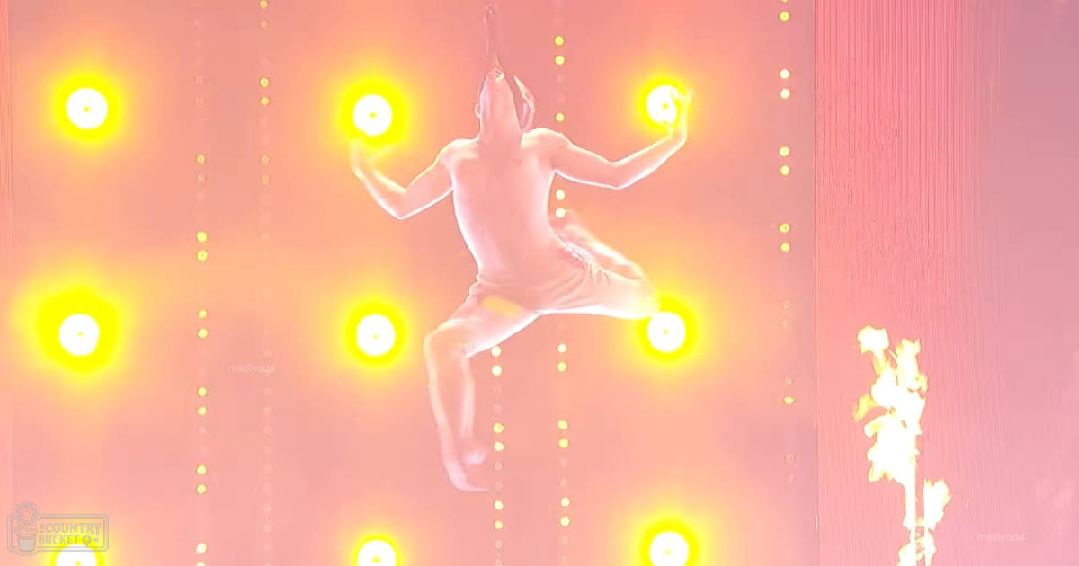 Young adult male performer in a smooth synthetic leotard, suspended by ropes with glowing discs on an indoor circus stage under warm lighting.