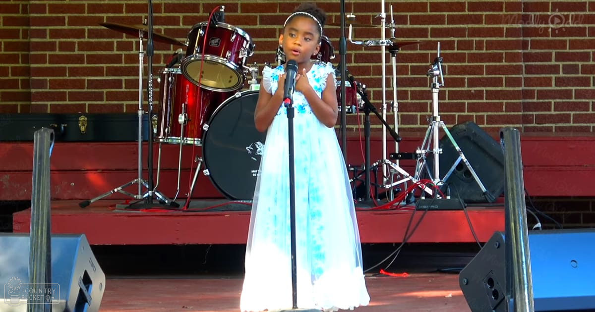 A young female child, approximately 5-8 years old, performs on stage at an outdoor event wearing a light blue dress with white accents and short sleeves, accompanied by a drum set in the background.
