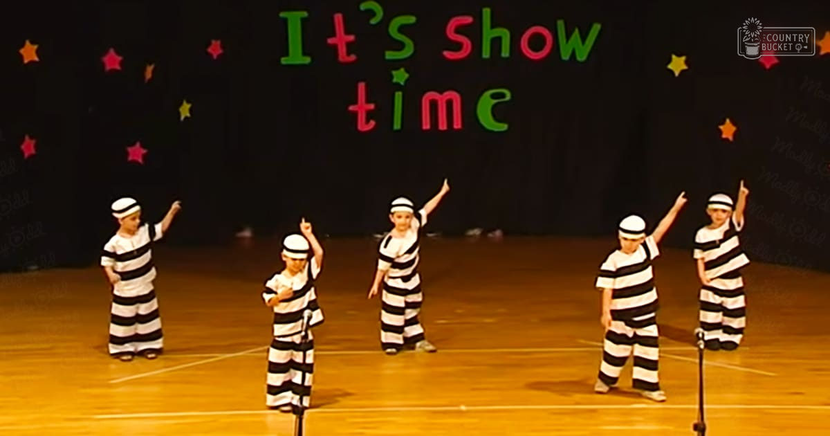 Children performing in a school setting, wearing black and white striped prison uniforms, barefoot or with socks, on stage with 'It's Showtime' text and star decorations.