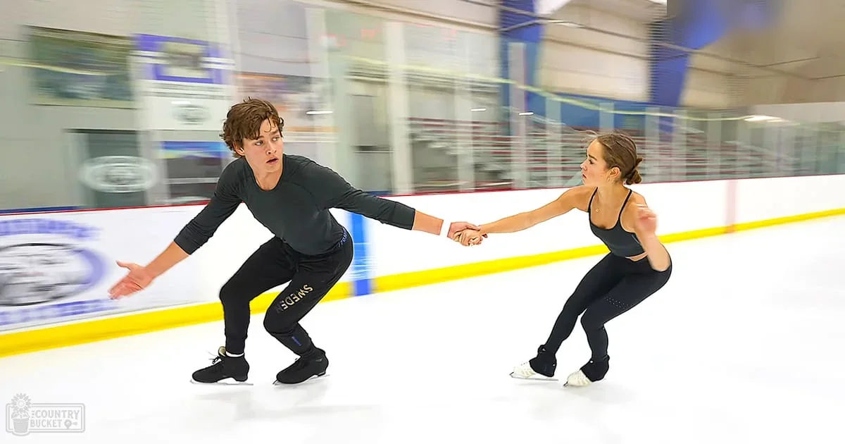A young couple skating together at an indoor ice rink with yellow boundary lines, evenly lit under artificial lighting.