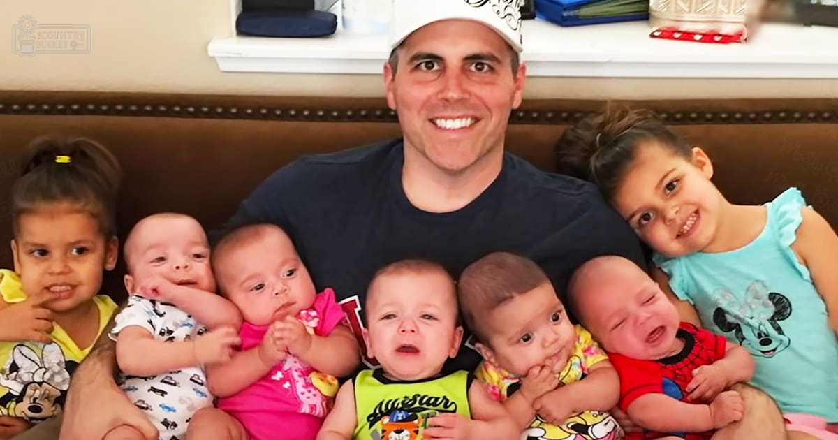 A middle-aged man indoors, surrounded by infants in colorful outfits; brown leather couch with decorative stitching and a white shelf holding various items. Text on clothing: 'Allstar'.